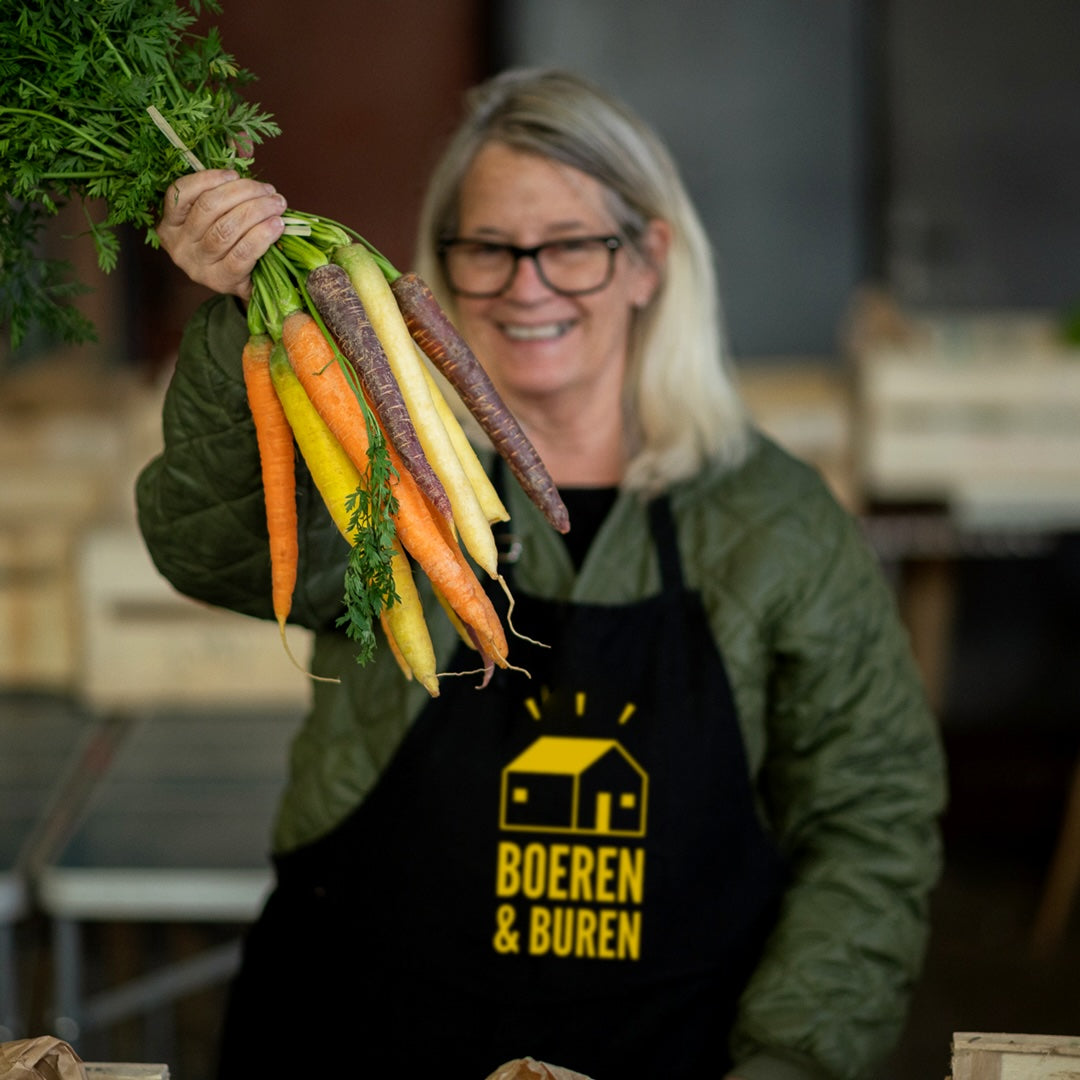 glimlachende vrouw met Boeren en Buren schort houdt een bos gekleurde wortels vast