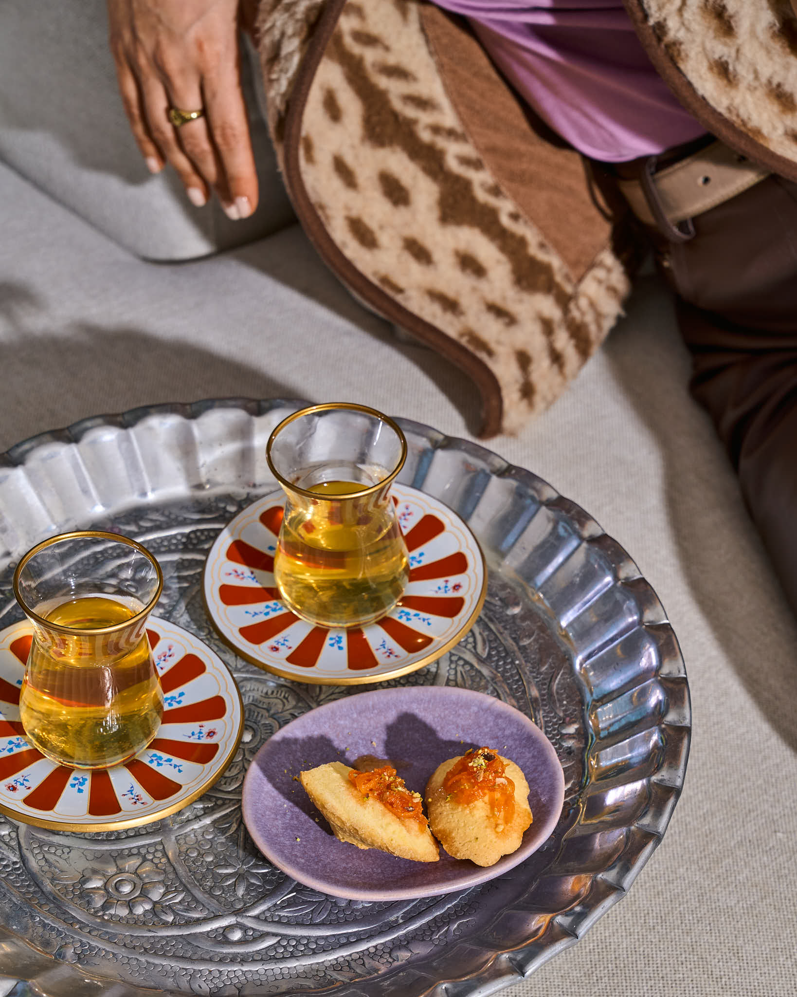Een zilveren dienblad met Turkse theeglazen en Madeleines op een zitbank met daarnaast het silhouet van een vrouw